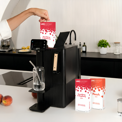 A black water dispenser. A hand is inserting a Strawberry and Raspberries Zero flavor packet into the machine. On the adjustable tray is a glass of water with a straw. To the right of the machine are two packages: Strawberry and Raspberries Zero and Langwater Peach Zero. Kitchen setting.
