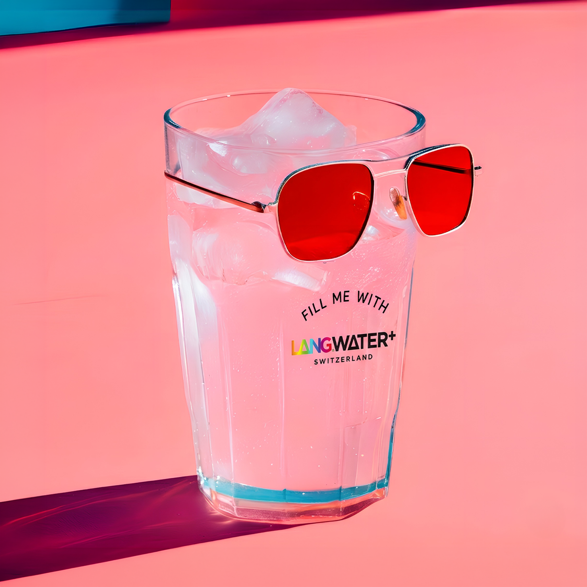 A clear glass filled with water and ice, with a pair of sunglasses resting on top and the colorful phrase Fill me with Langwater Switzerland in the center of the glass, against a red background.